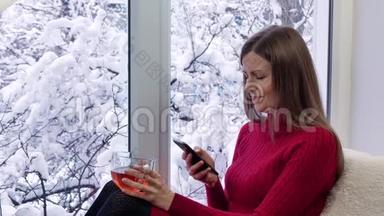 穿着红裙子的漂亮女孩坐在窗台上，喝茶，用智能手机。 冬天在外面。